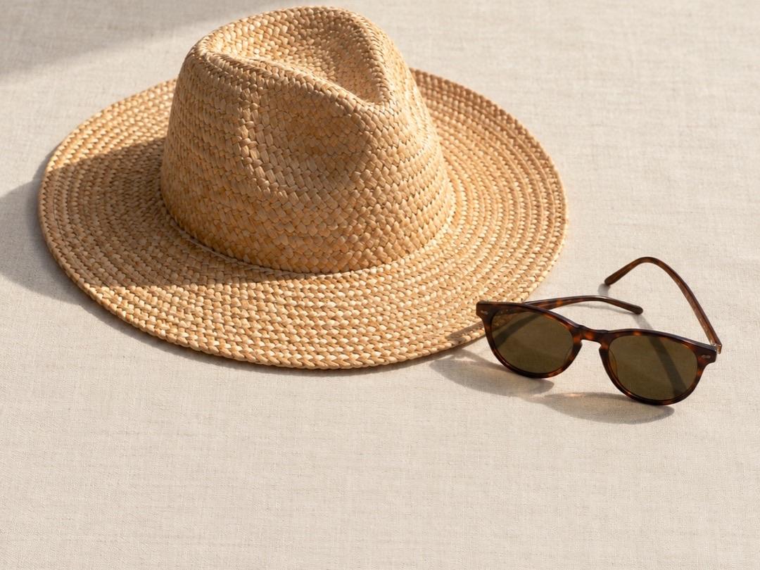 Woven straw sun hat and sunglasses on a light linen cloth in bright sunlight