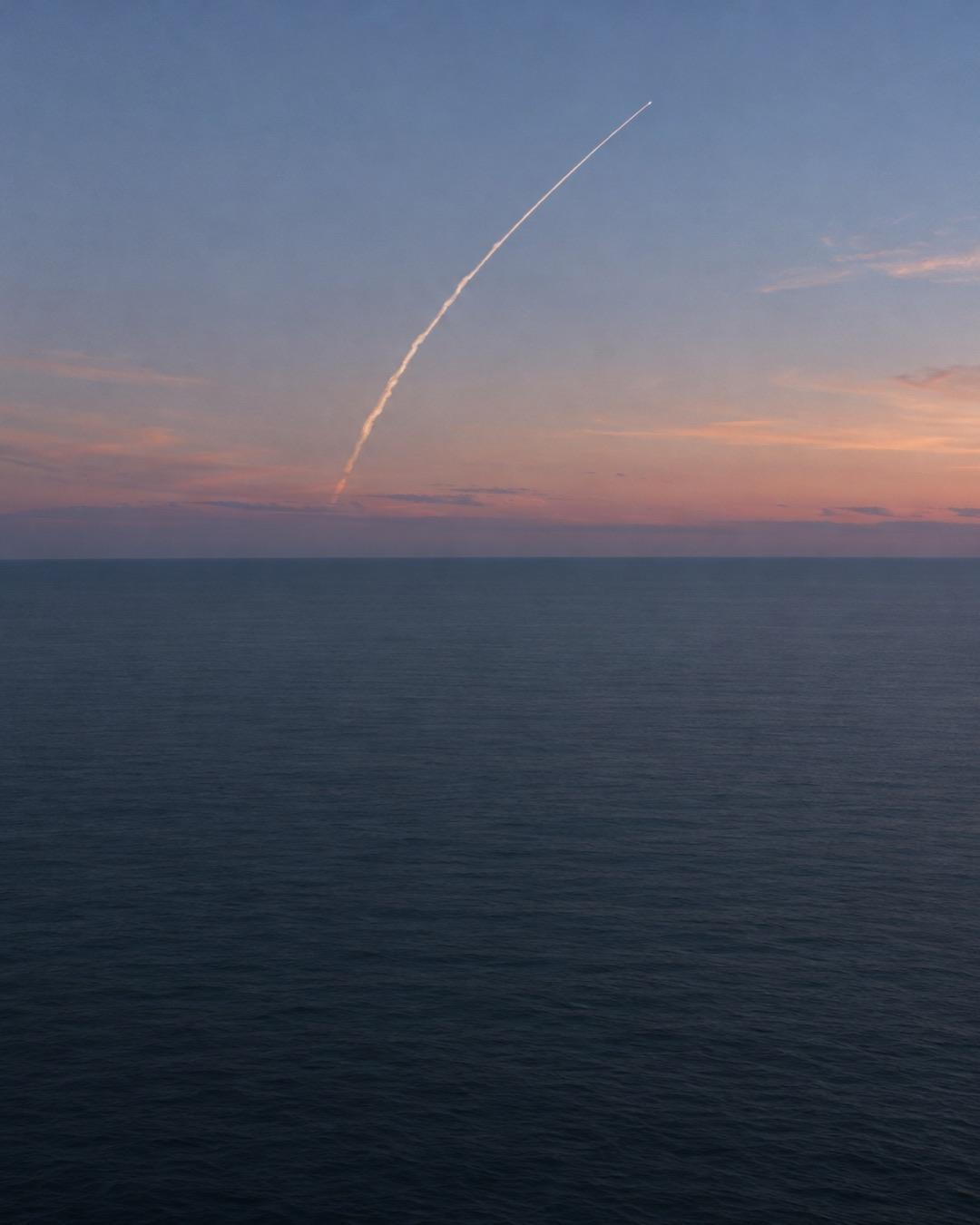 Thin rocket exhaust trail curving across an evening sky over a calm sea